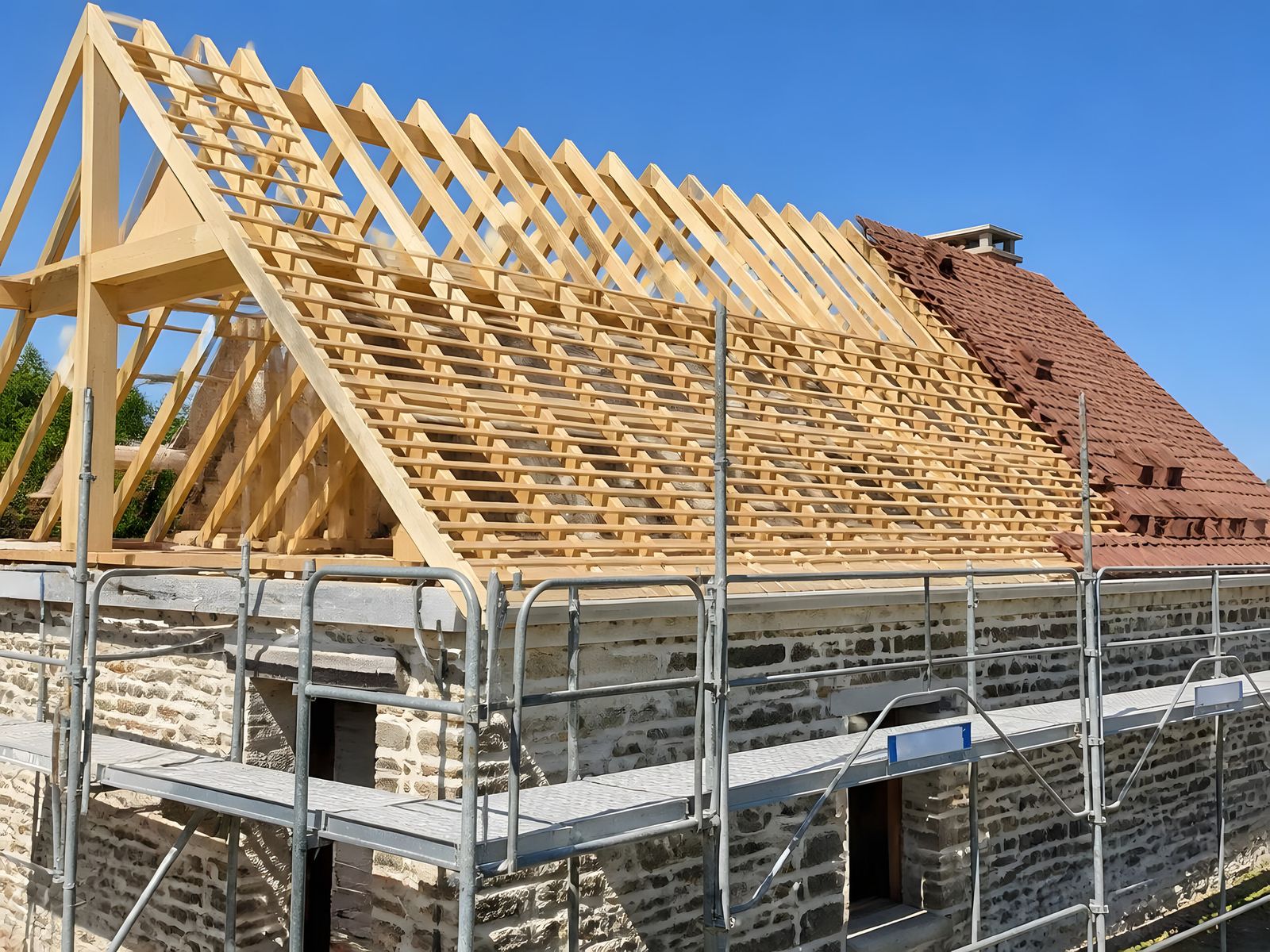 Bâtisse en pierre sous échafaudage : ferme, chevrons et liteaux neufs en bois blond, vieilles tuiles encore en place à droite