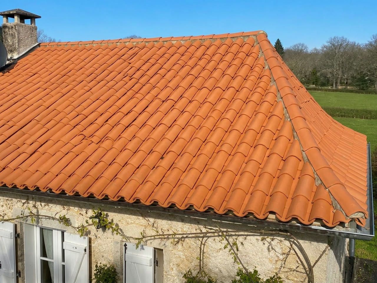 Toiture à croupe en tuiles canal orangées, arêtier au mortier et gouttière en zinc, sur une maison en pierre